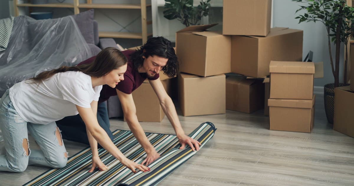 Una pareja extiende la alfombra en el salón de su piso nuevo, entre cajas de mudanza