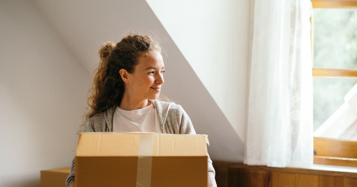 Una joven sonríe junto a la ventana de su primera casa, con una caja de mudanza delante