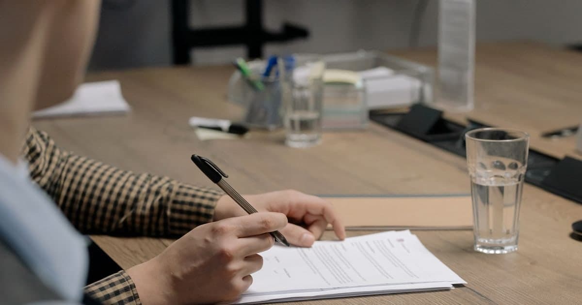 Una persona se dispone a firmar unos papeles con bolígrafo sobre la mesa de madera de una oficina, con un vaso de agua al lado