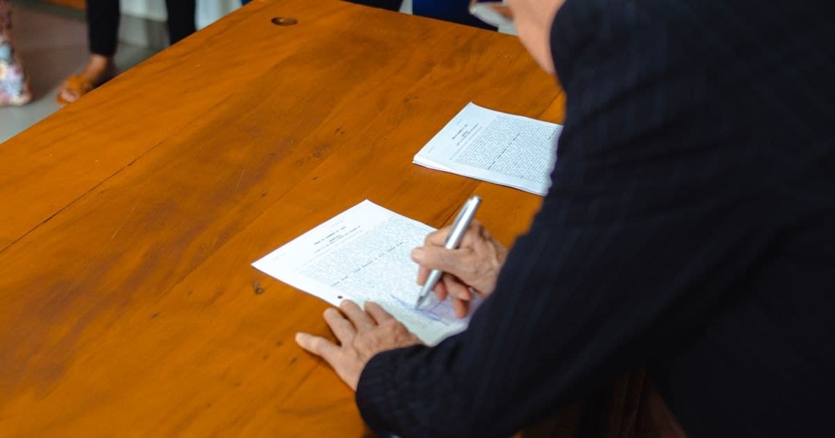 Una persona de traje firma unos papeles con bolígrafo sobre una mesa grande de madera