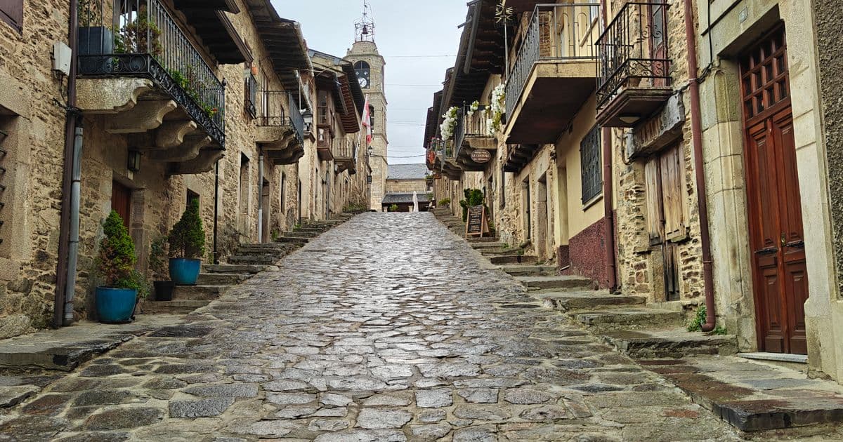 Calle empedrada de un pueblo de Castilla y León entre casas de piedra con galerías de madera y balcones de forja, con la torre del reloj al fondo