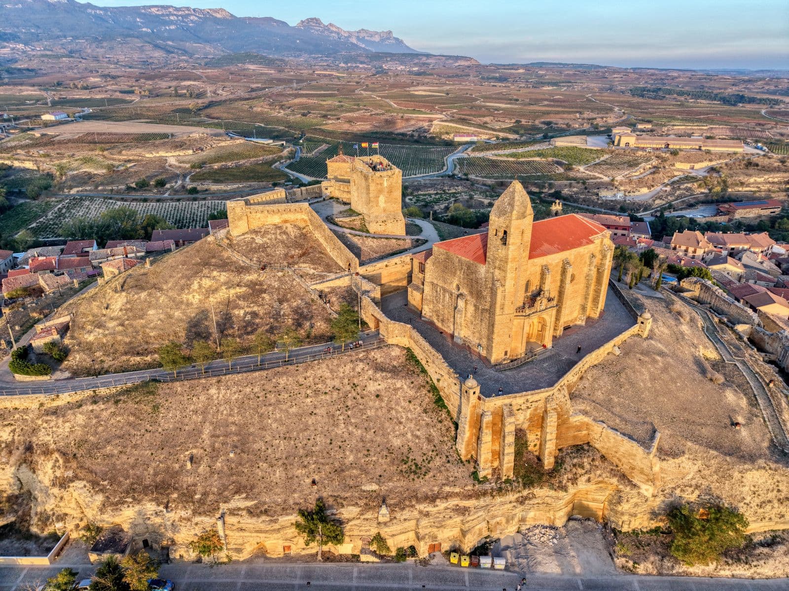Vista aérea de un pueblo riojano en alto con iglesia y murallas de castillo, casas de tejado rojo y viñedos alrededor