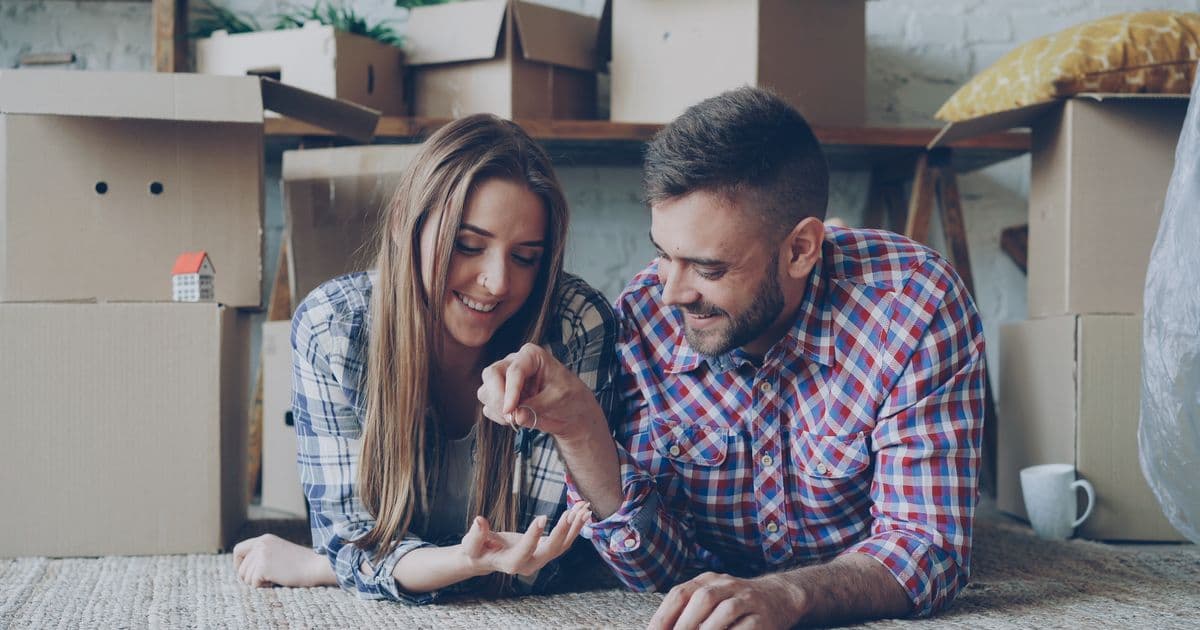 Pareja joven sonriendo rodeada de cajas de mudanza en su primera vivienda