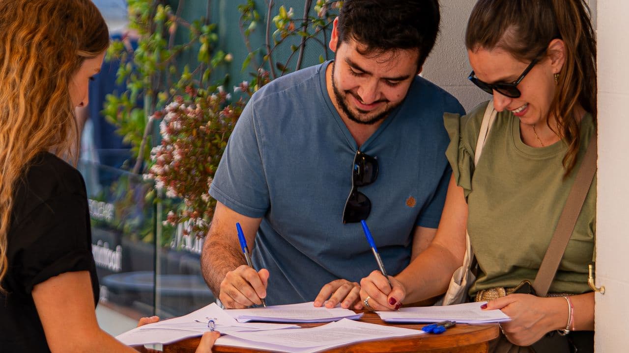 Una pareja firma documentos sobre una mesa, de pie y sonriendo